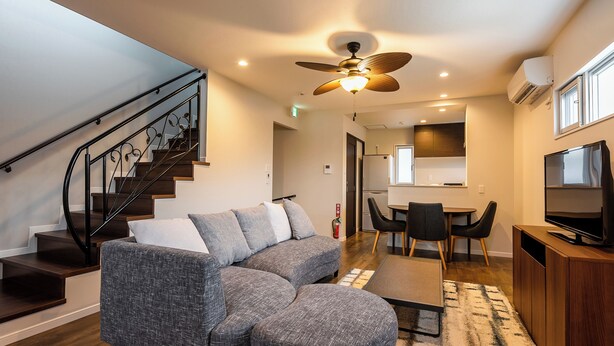 A modern living room with a grey sectional sofa, a small ottoman, a dining area with a table and chairs, a ceiling fan, and a TV on a wooden cabinet. - Tsukasa Condo Chatan Araha (Chatan)