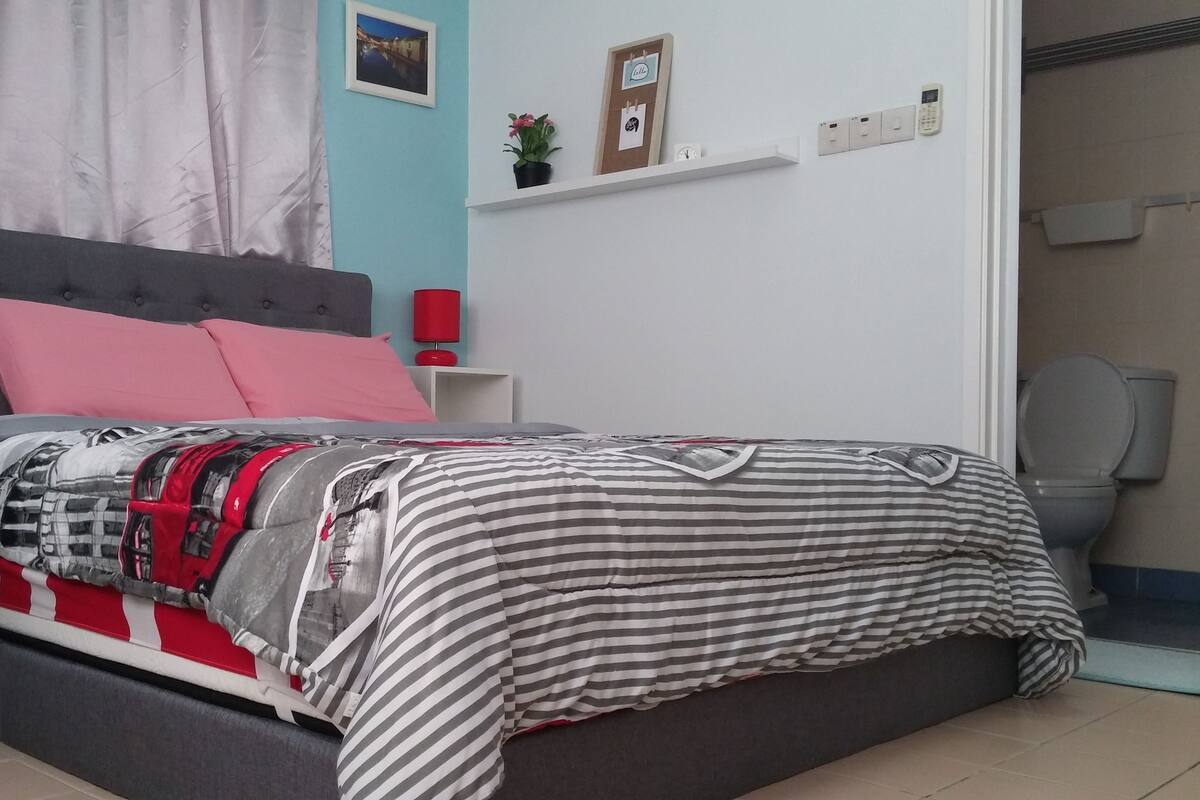 A bedroom with a bed, pillows, a nightstand, a red lamp, a wall clock, and a small potted plant.