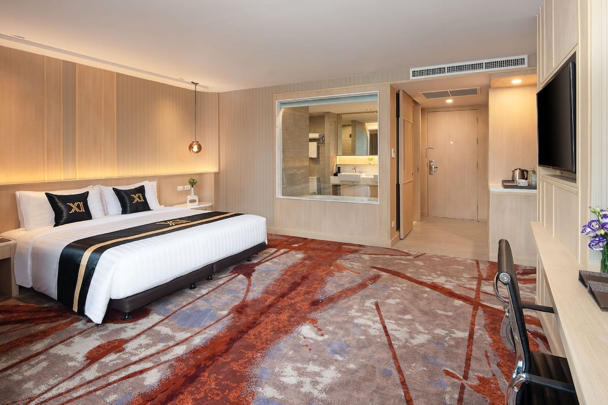 A modern hotel room with a large bed, a desk, a TV, and a bathroom visible through a glass door.