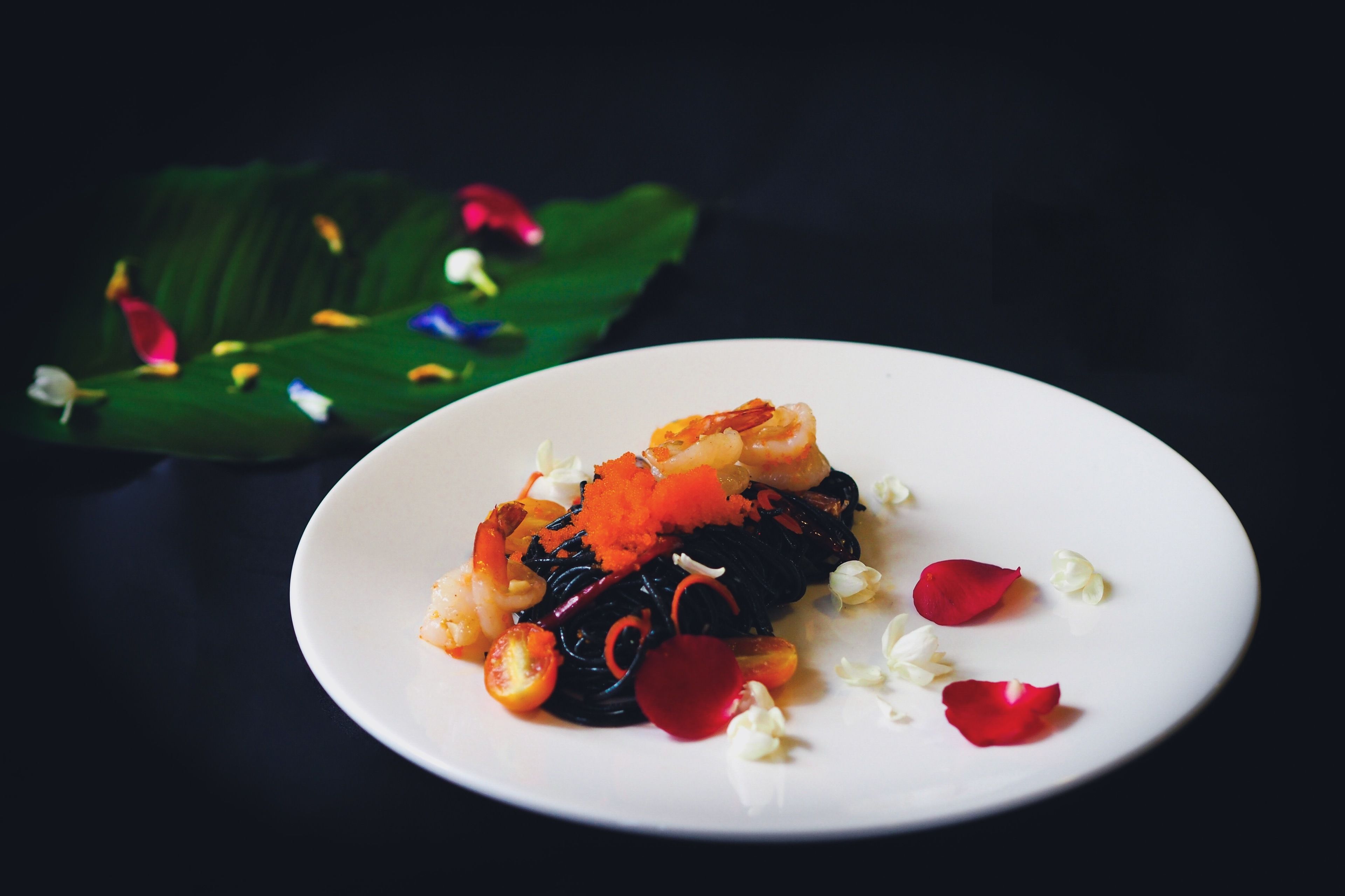 A plate of food with shrimp, vegetables, and garnishes, placed on a dark surface with a green leaf and colorful decorations in the background.