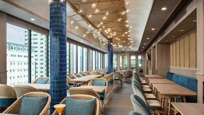 Een modern restaurant met hoge plafonds, rieten meubels en grote ramen met uitzicht over de stad.