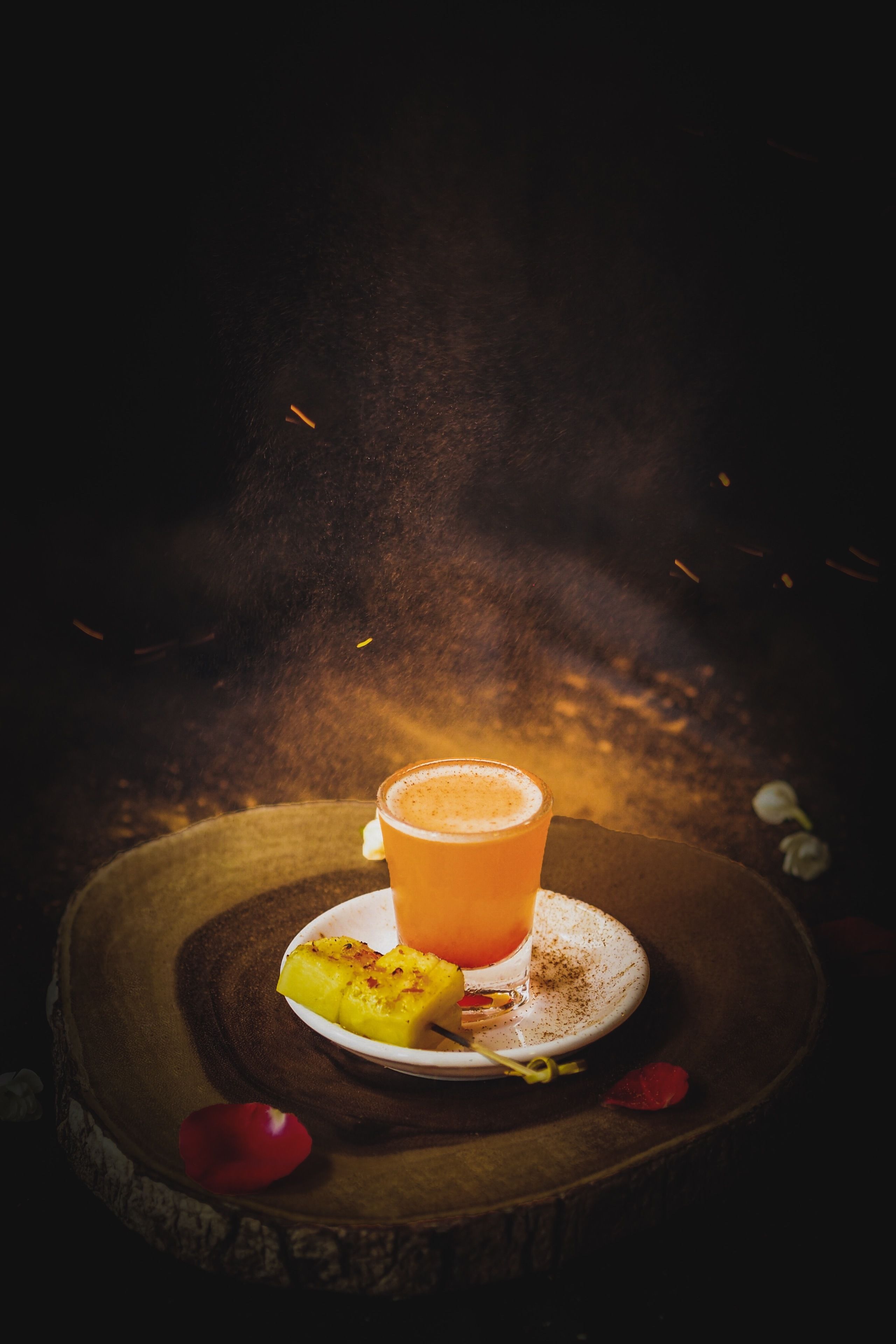A glass of orange juice on a wooden table with a slice of pineapple and a flower petal.