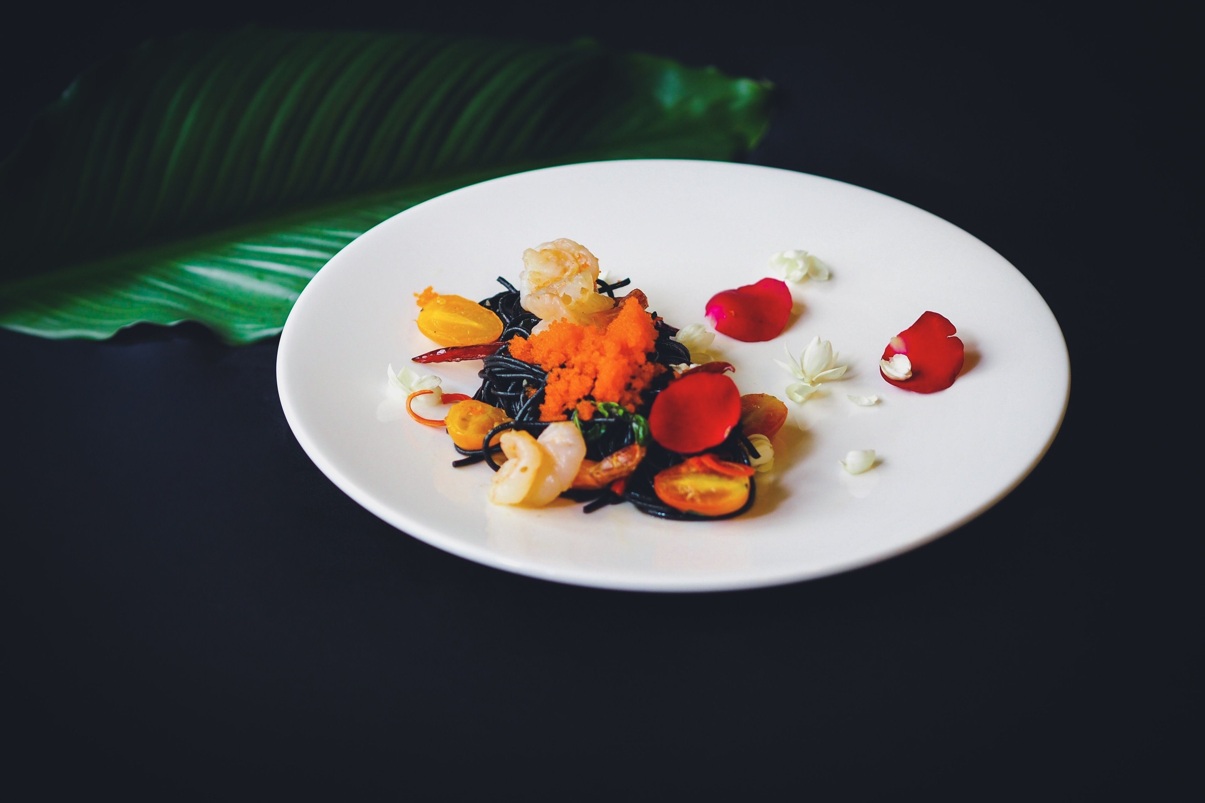 A white plate with a variety of food items including shrimp, vegetables, and garnishes, set against a dark background with a green leaf.