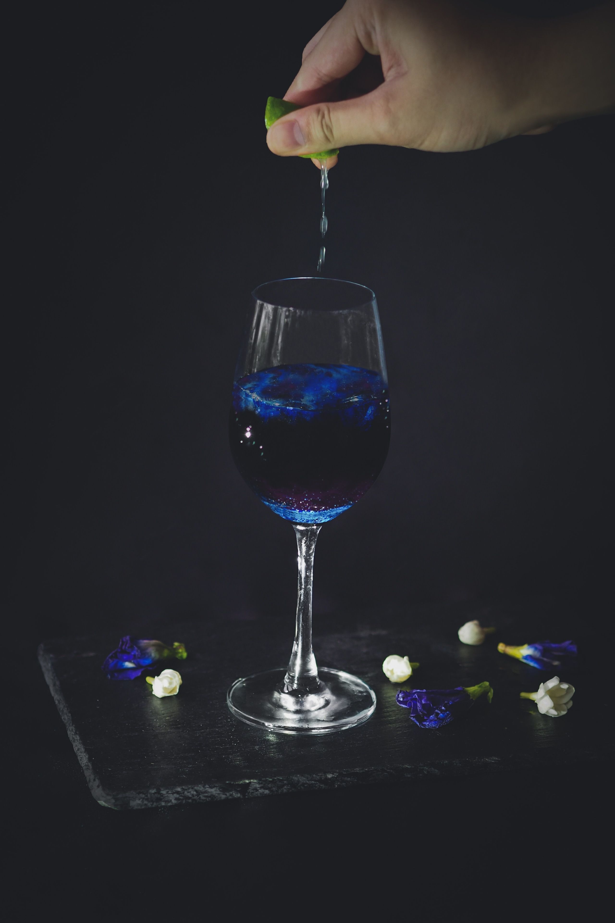A glass of blue liquid with a hand pouring more liquid into it.