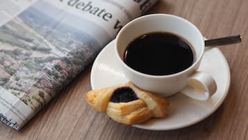 A cup of coffee on a saucer with a spoon, a pastry, and a newspaper on a wooden table.