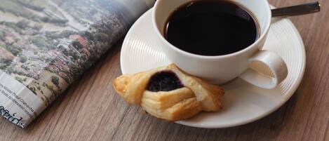 A cup of coffee on a saucer with a spoon, a pastry, and a newspaper on a wooden table.