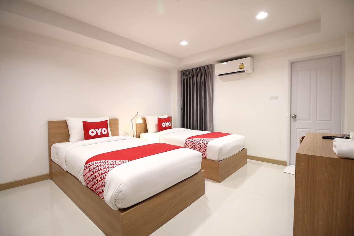 Une chambre d’hôtel avec deux lits, chacun agrémenté de draps blancs et de touches de couleur rouge, une tête de lit en bois, un bureau avec une chaise et un appareil de climatisation fixé au mur.