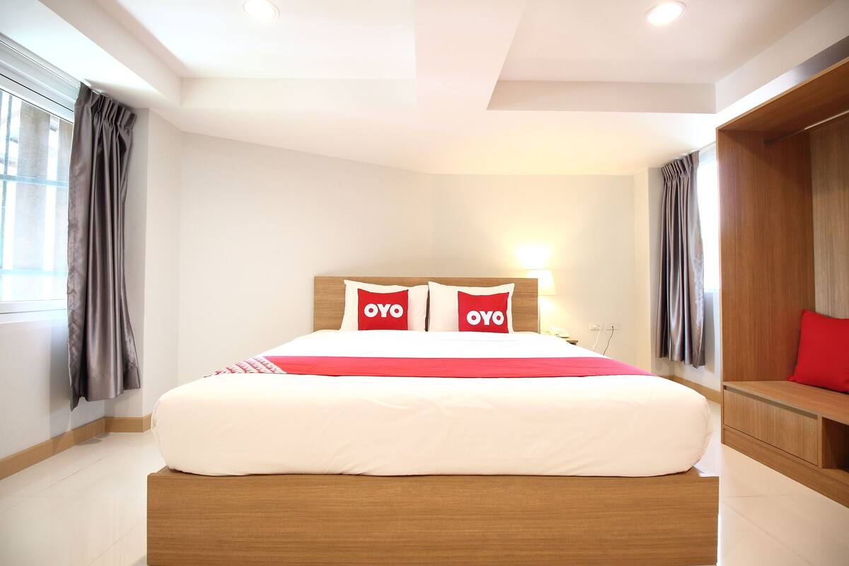 Une chambre d’hôtel moderne avec un grand lit, deux oreillers portant la marque « OYO », une tête de lit en bois et un banc intégré avec un coussin rouge.