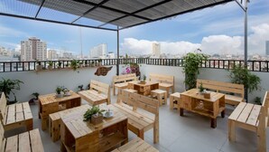 An outdoor seating area with wooden tables and benches, surrounded by potted plants and overlooking a cityscape.
