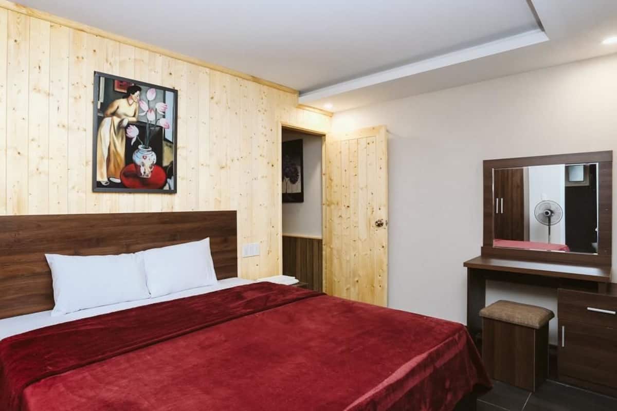 A bedroom with a wooden headboard, a red bedspread, a wooden wardrobe, a mirror, and a painting on the wall.
