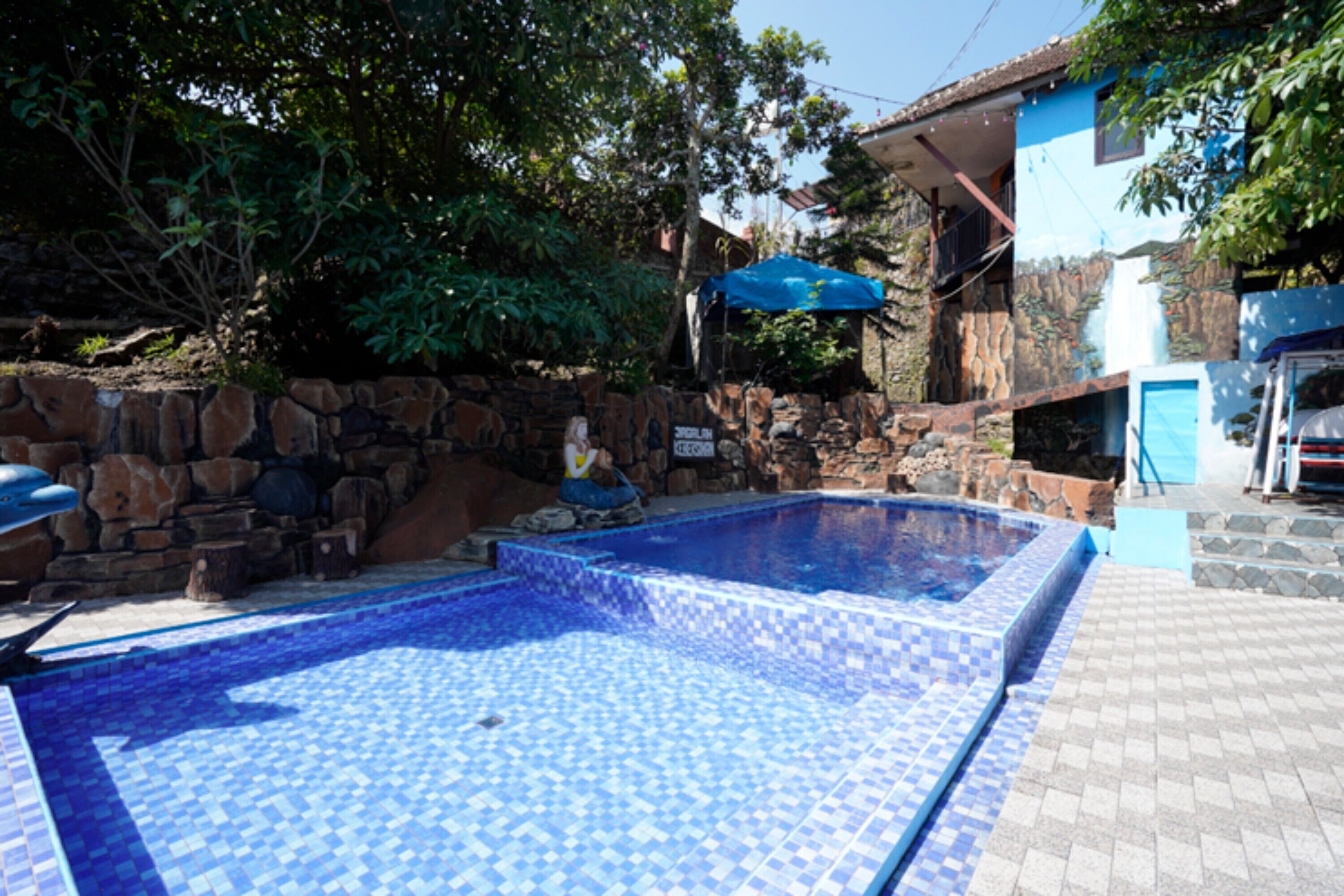 A swimming pool with blue mosaic tiles, surrounded by a stone wall and a blue umbrella.