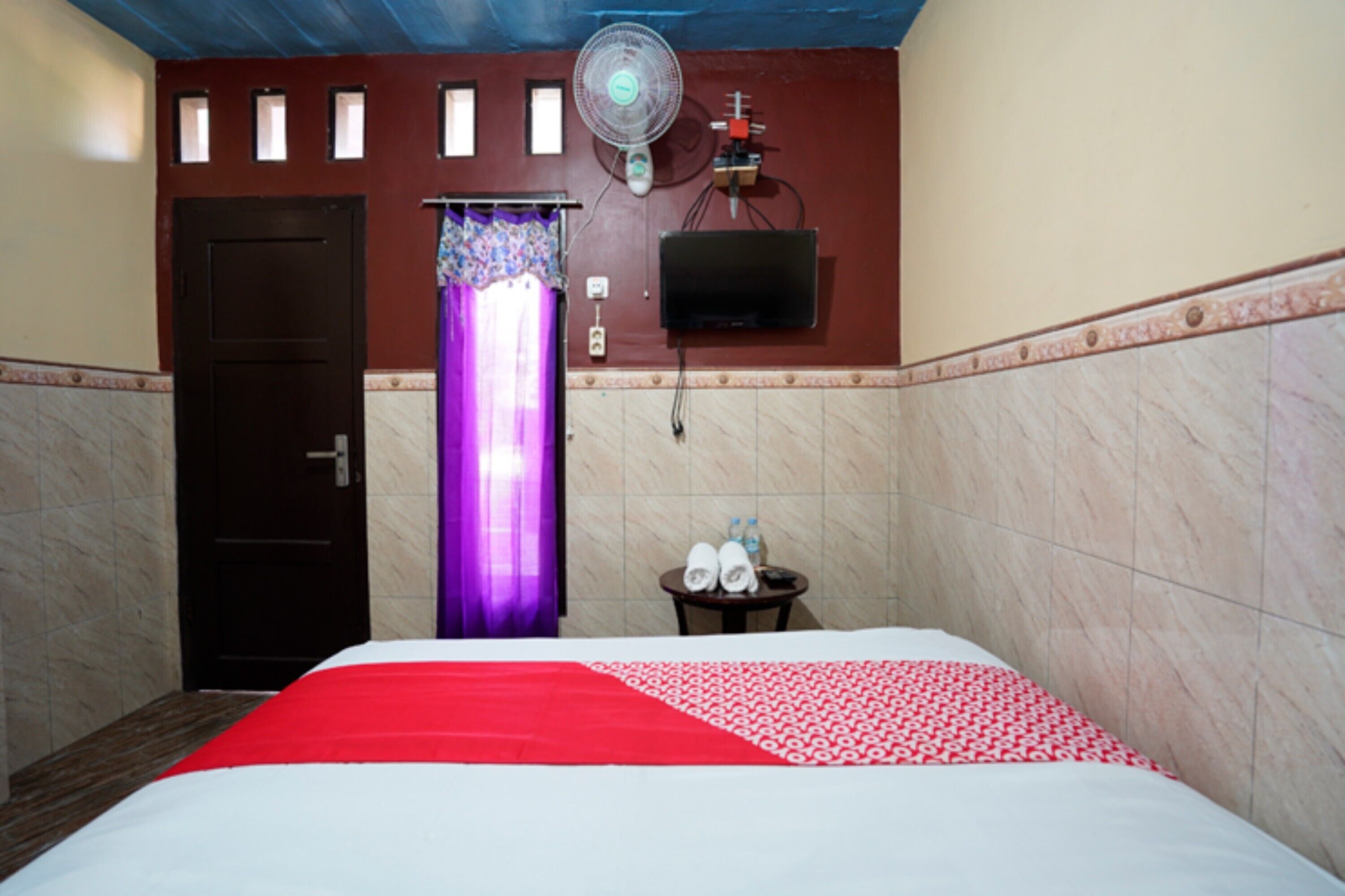 A hotel room with a bed, a door, a wall-mounted TV, a fan, and a small table with towels.