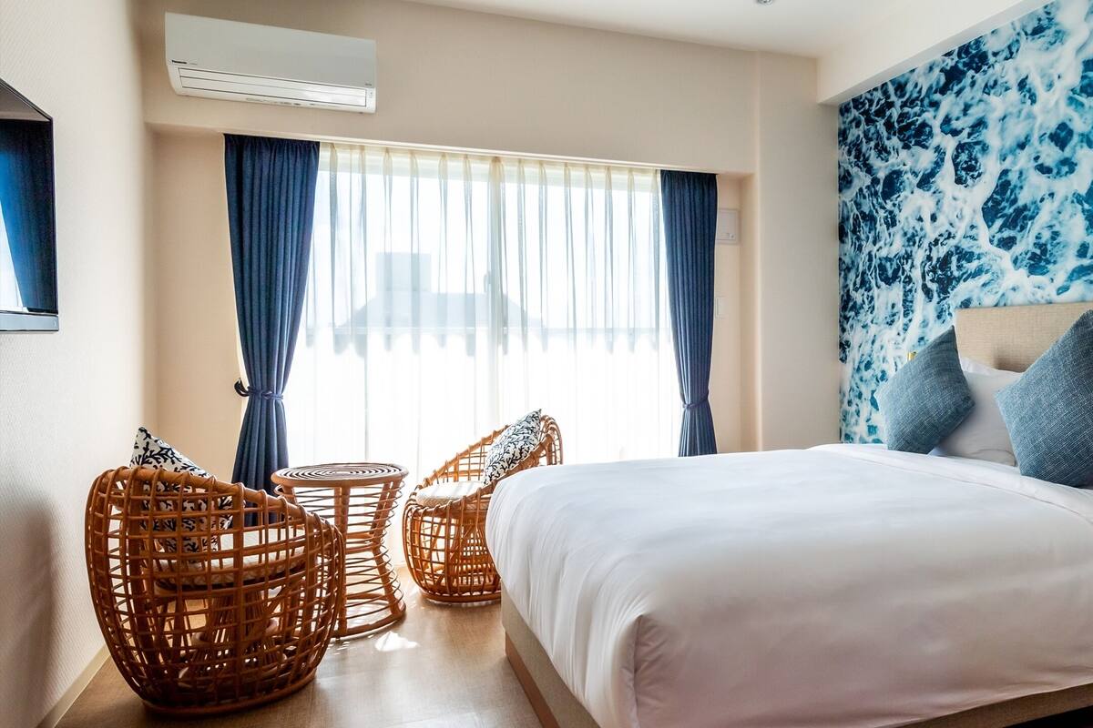 A bedroom with a bed, two wicker chairs, a small table, a nightstand, and a wall with a blue abstract pattern.