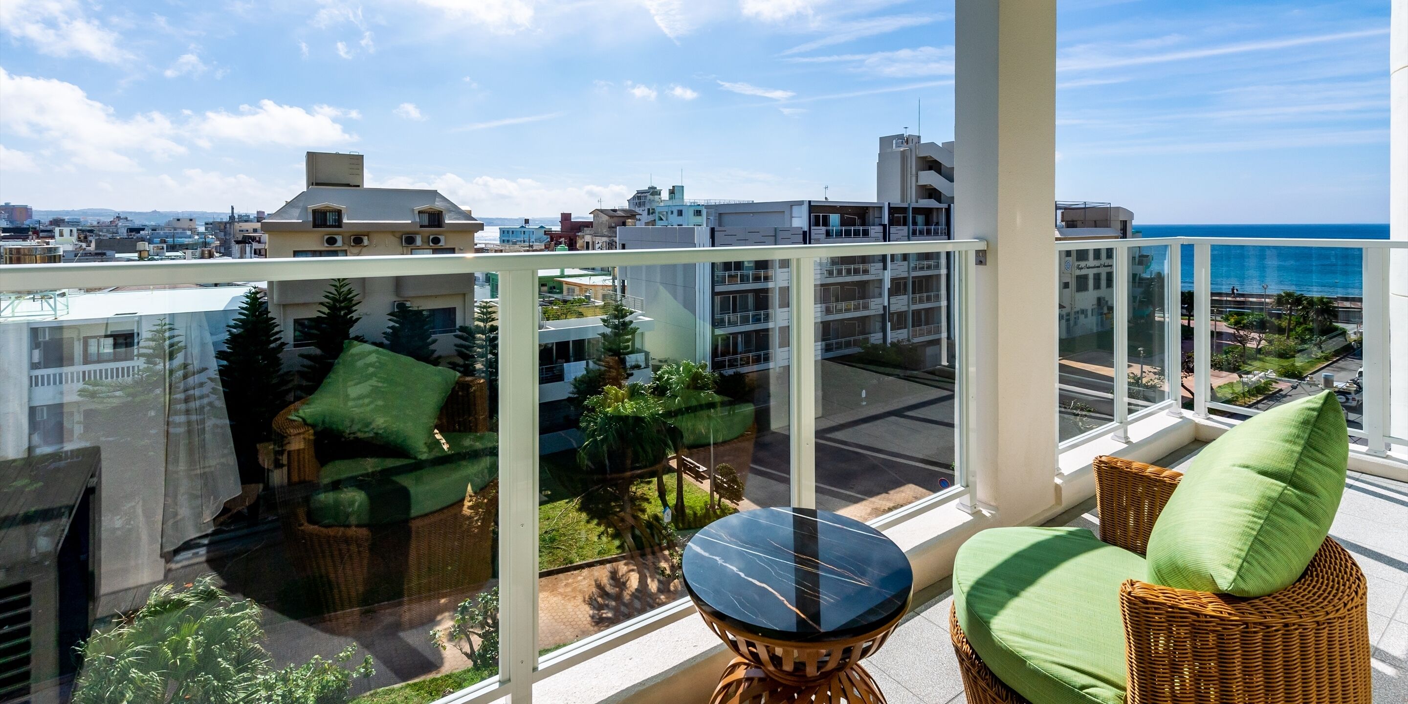 A balcony with wicker furniture, a glass table, and a view of the city and ocean.