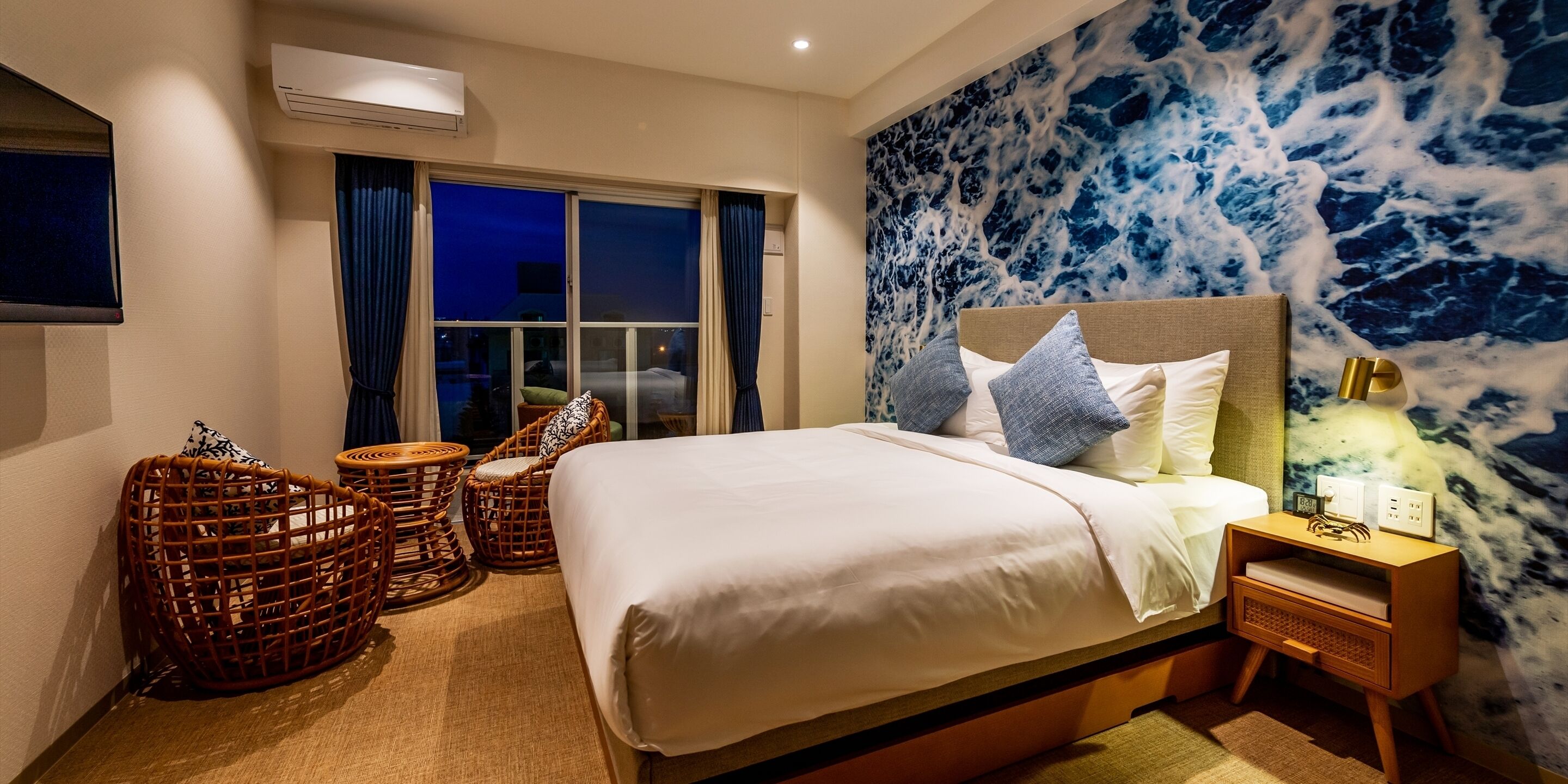 Quarto com uma cama grande, cadeiras de vime, uma mesa de cabeceira e um mural na parede.
