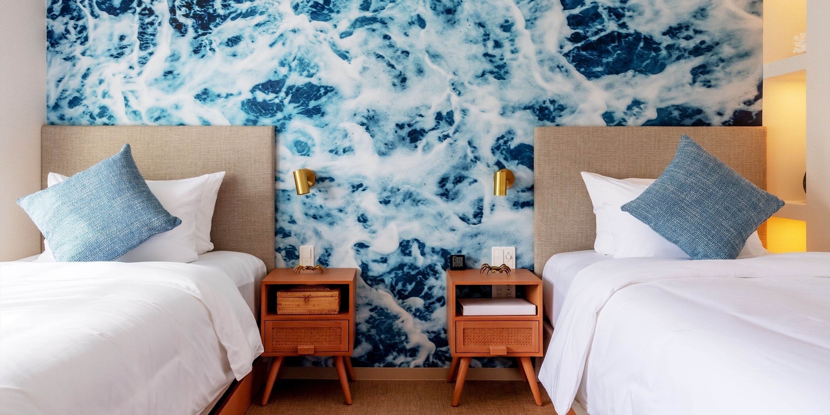 A hotel room with two beds, white bedding, blue pillows, wooden nightstands, and a wall mural of blue and white abstract patterns.