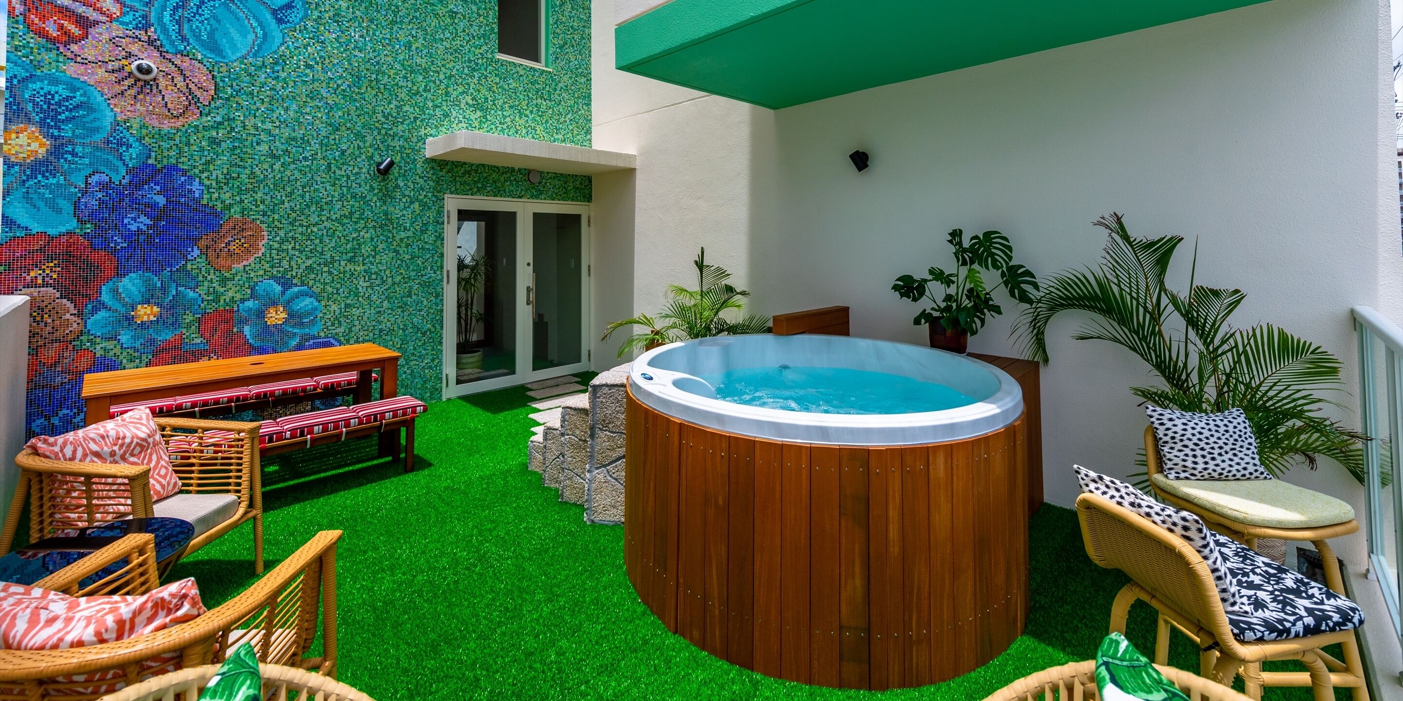 Un patio con césped artificial, un tinaco de madera caliente, mobiliario de mimbre y una pared de mosaicos.