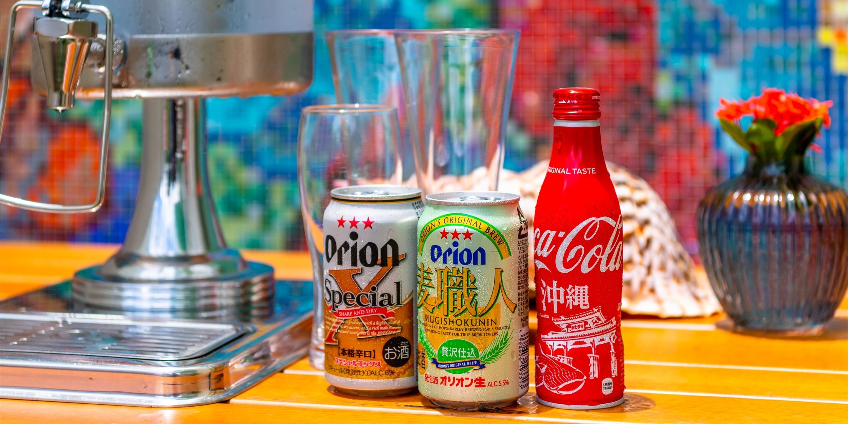 Una mesa con un dispensador de soda, dos latas de Orion Special, una botella de Coca-Cola y un florero con flores.