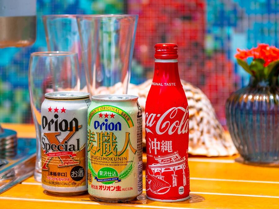 Una mesa con un dispensador de soda, dos latas de Orion Special, una botella de Coca-Cola y un florero con flores.