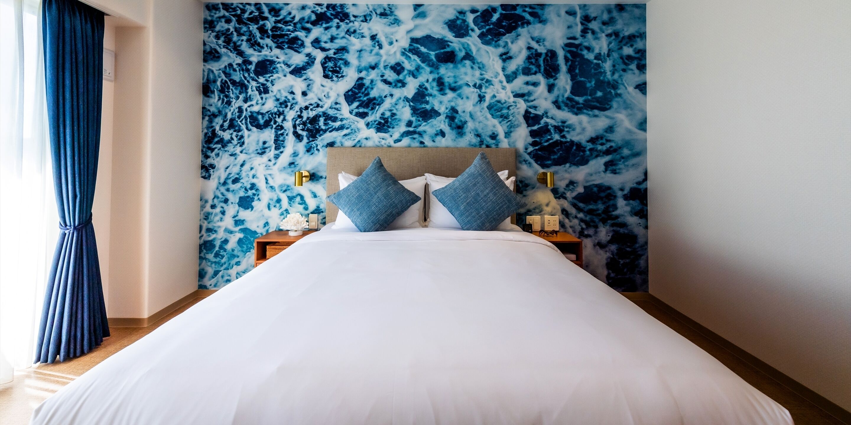 A hotel room with a bed, a headboard, two pillows, a nightstand, and a wall with a blue and white pattern.