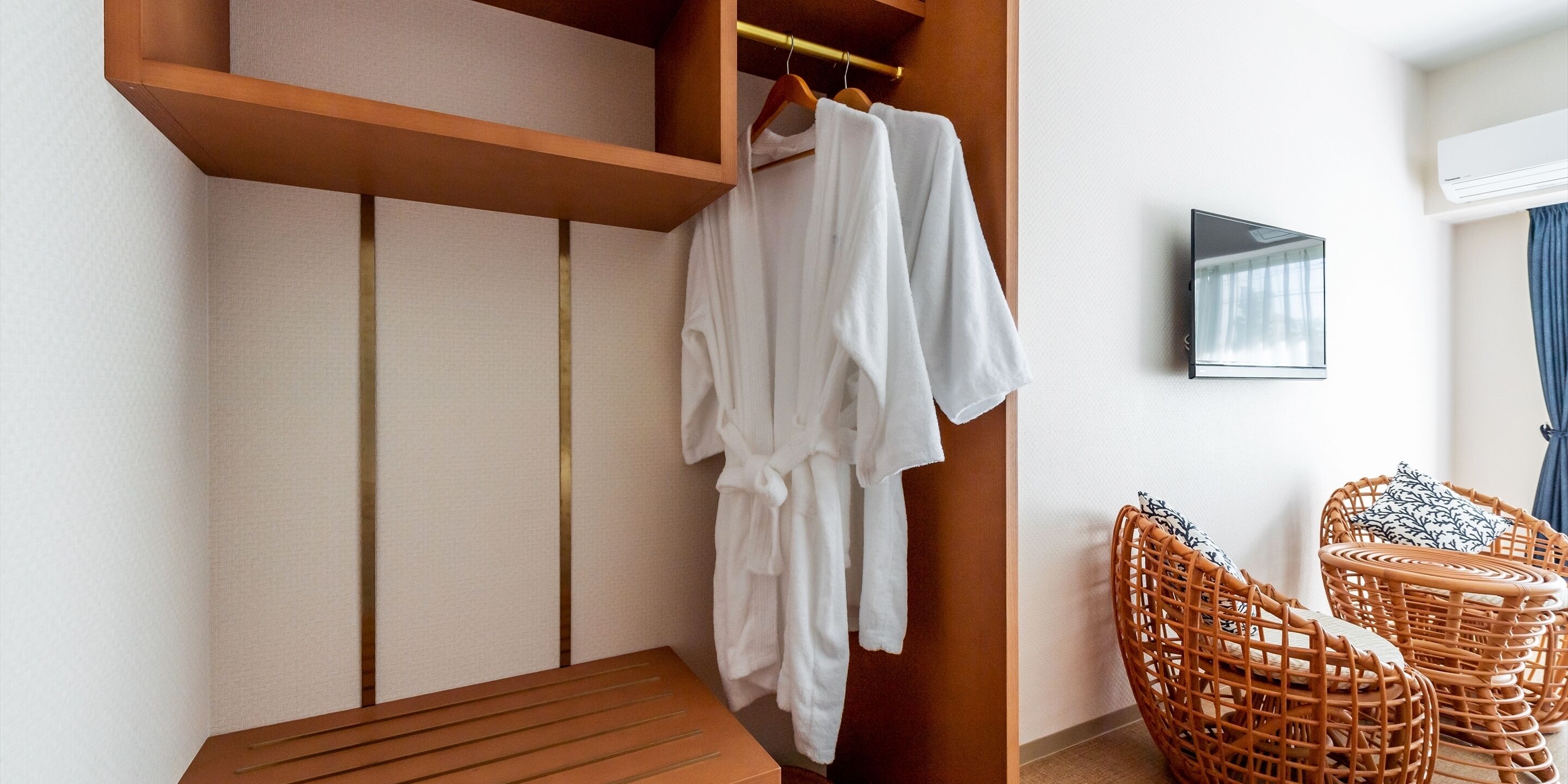 Una habitación con un banco de madera, una bata blanca colgada, sillas de mimbre y un televisor montado.