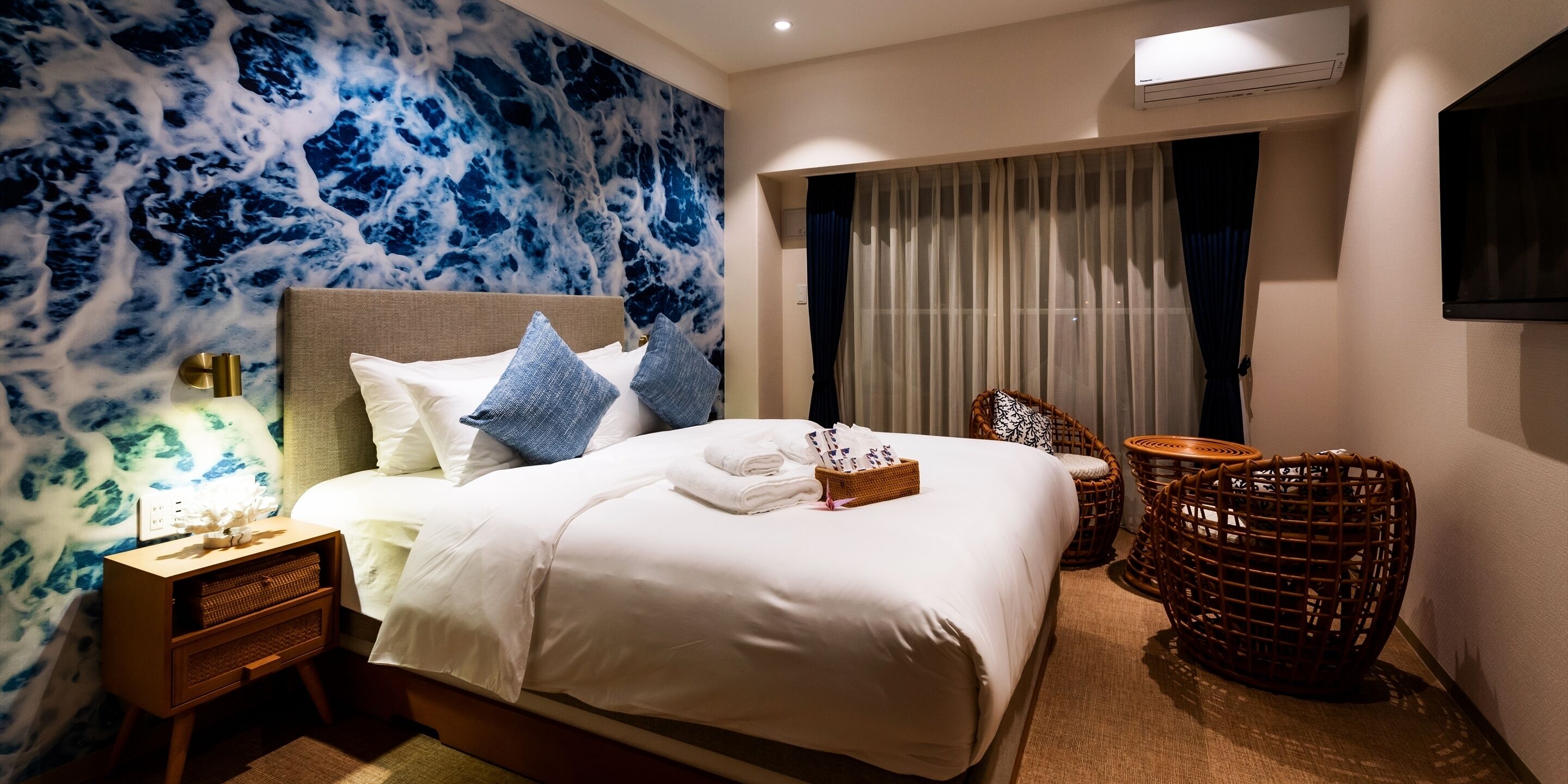 A neatly made bed with white linens and blue pillows, a bedside table with a lamp, and a wall with a blue and white abstract pattern.