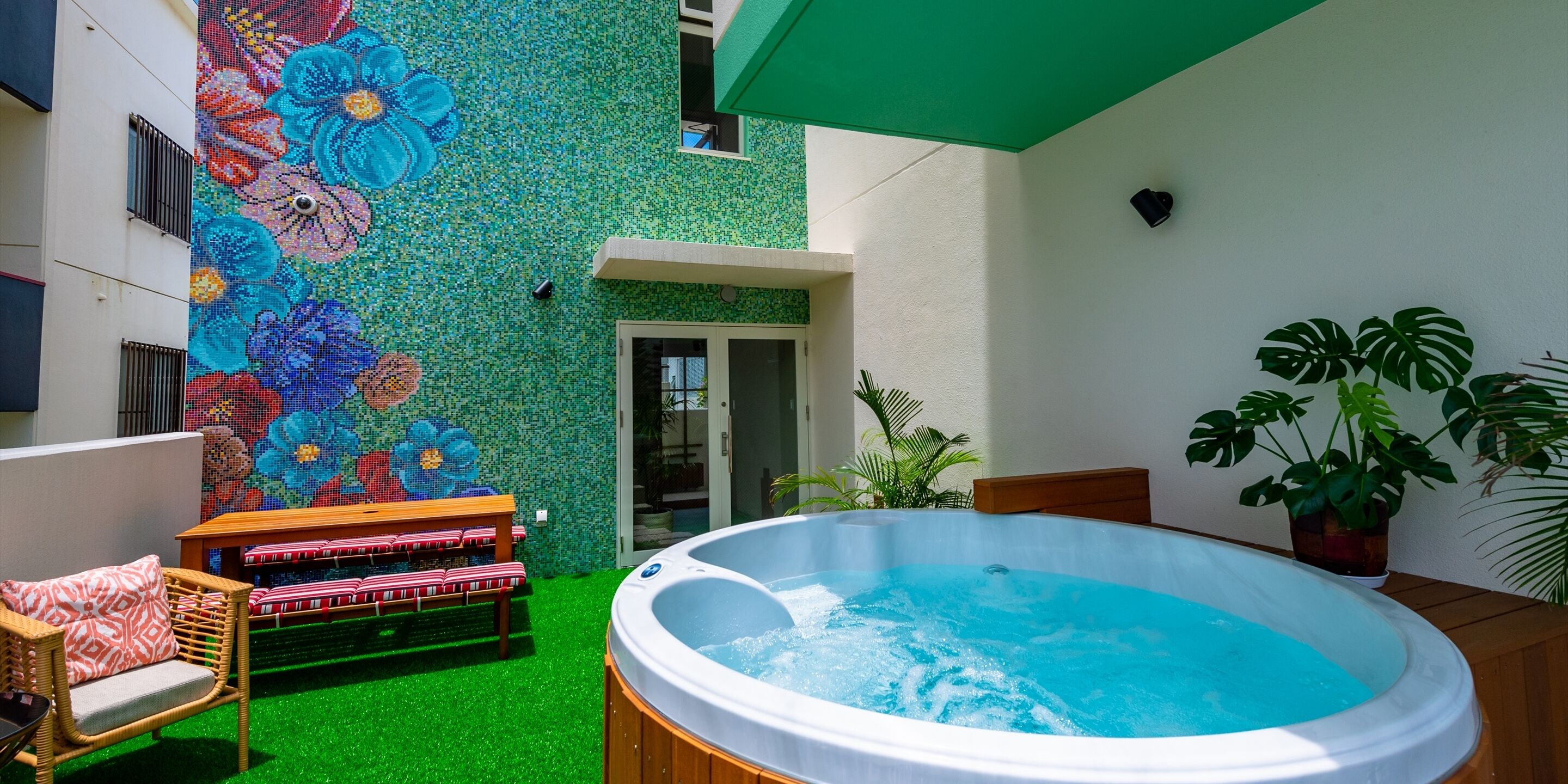 A hot tub in a courtyard with a mosaic wall, a wooden bench, and potted plants.
