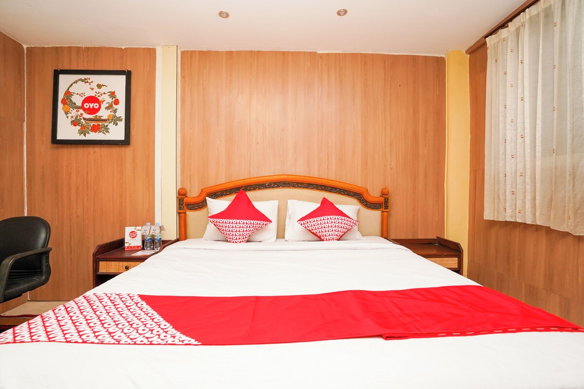 Une chambre d’hôtel avec une tête de lit en bois, un lit avec une literie blanche et rouge, un plaid à motifs rouges et blancs, un tableau encadré au mur et une chaise.