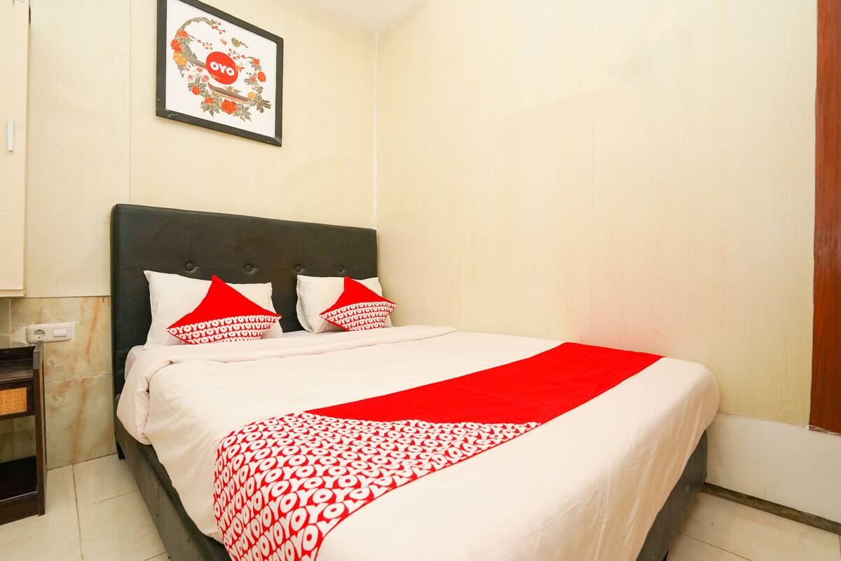 Una cama bien hecha con una colcha blanca y roja estampada, dos almohadas rojas y una obra de arte floral enmarcada sobre la cama.
