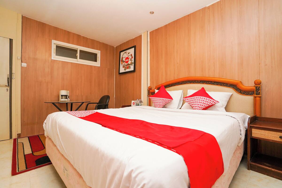 Habitación de hotel con pared de madera, cama con ropa de cama blanca y detalles rojos, una mesita con una silla y un cuadro en la pared.