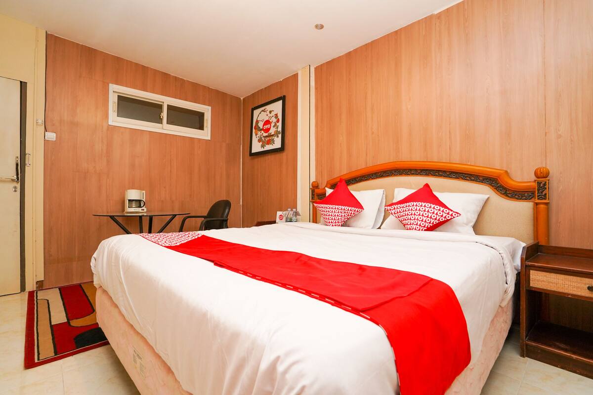 Habitación de hotel con pared de madera, cama con ropa de cama blanca y detalles rojos, una mesita con una silla y un cuadro en la pared.