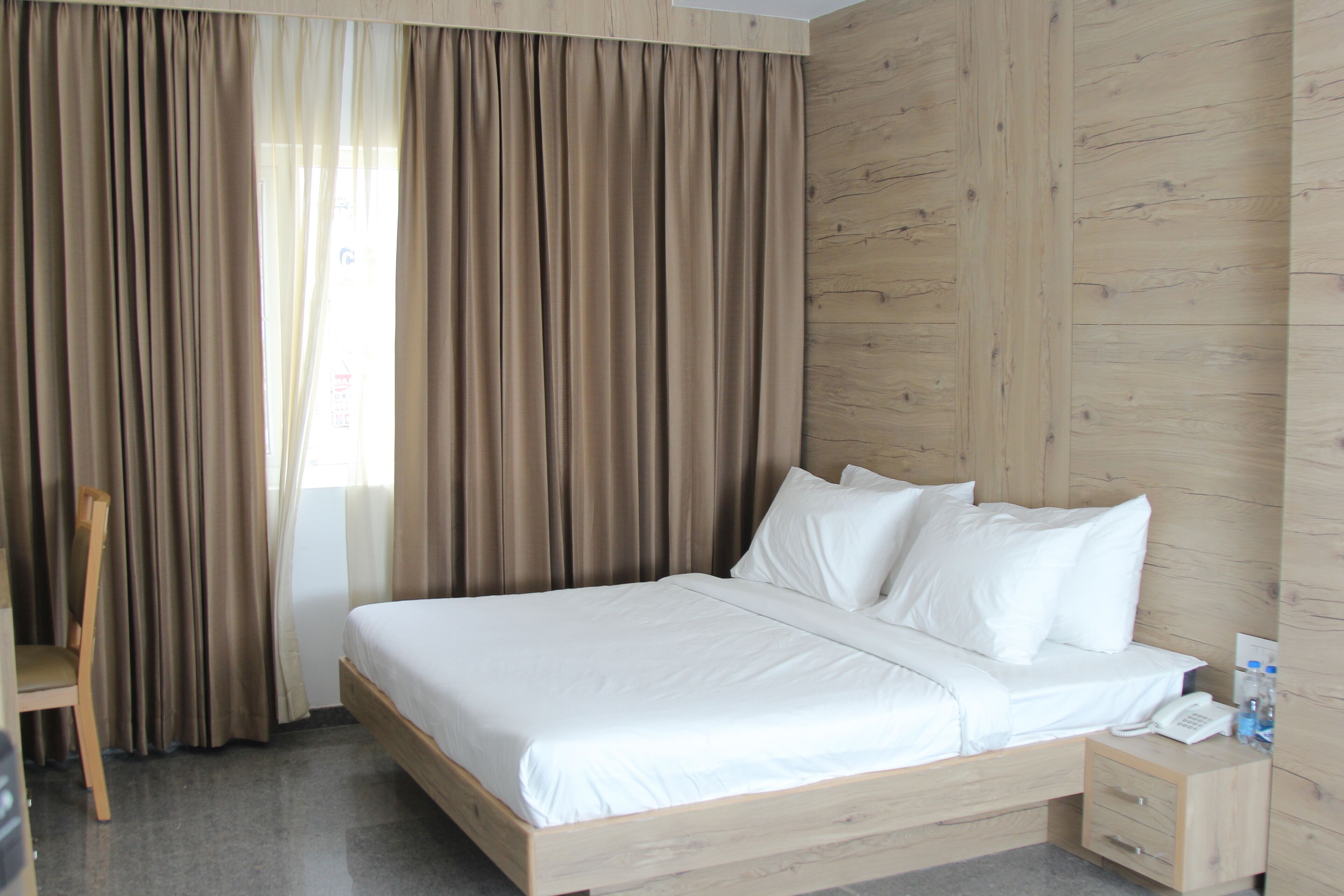Habitación de hotel con cama de madera, ropa de cama blanca, mesita de noche de madera con un teléfono y botellas de agua, una silla de madera y una ventana con cortinas.