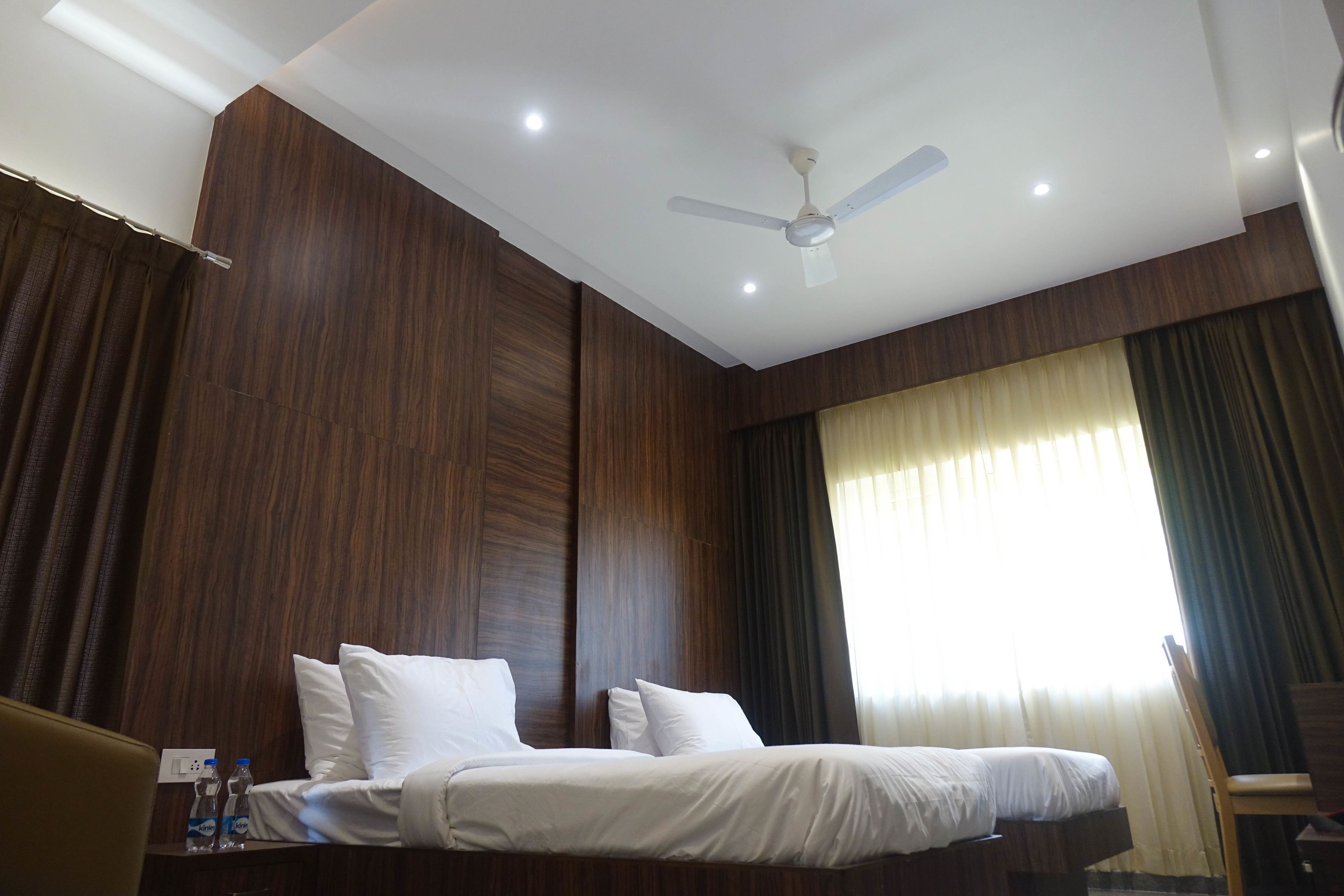 Una habitación de hotel con una cama, un ventilador de techo y una ventana con cortinas.