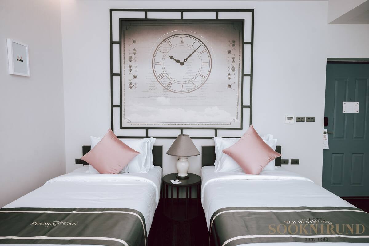 A hotel room with two beds, a large wall clock, and a small table with a lamp.