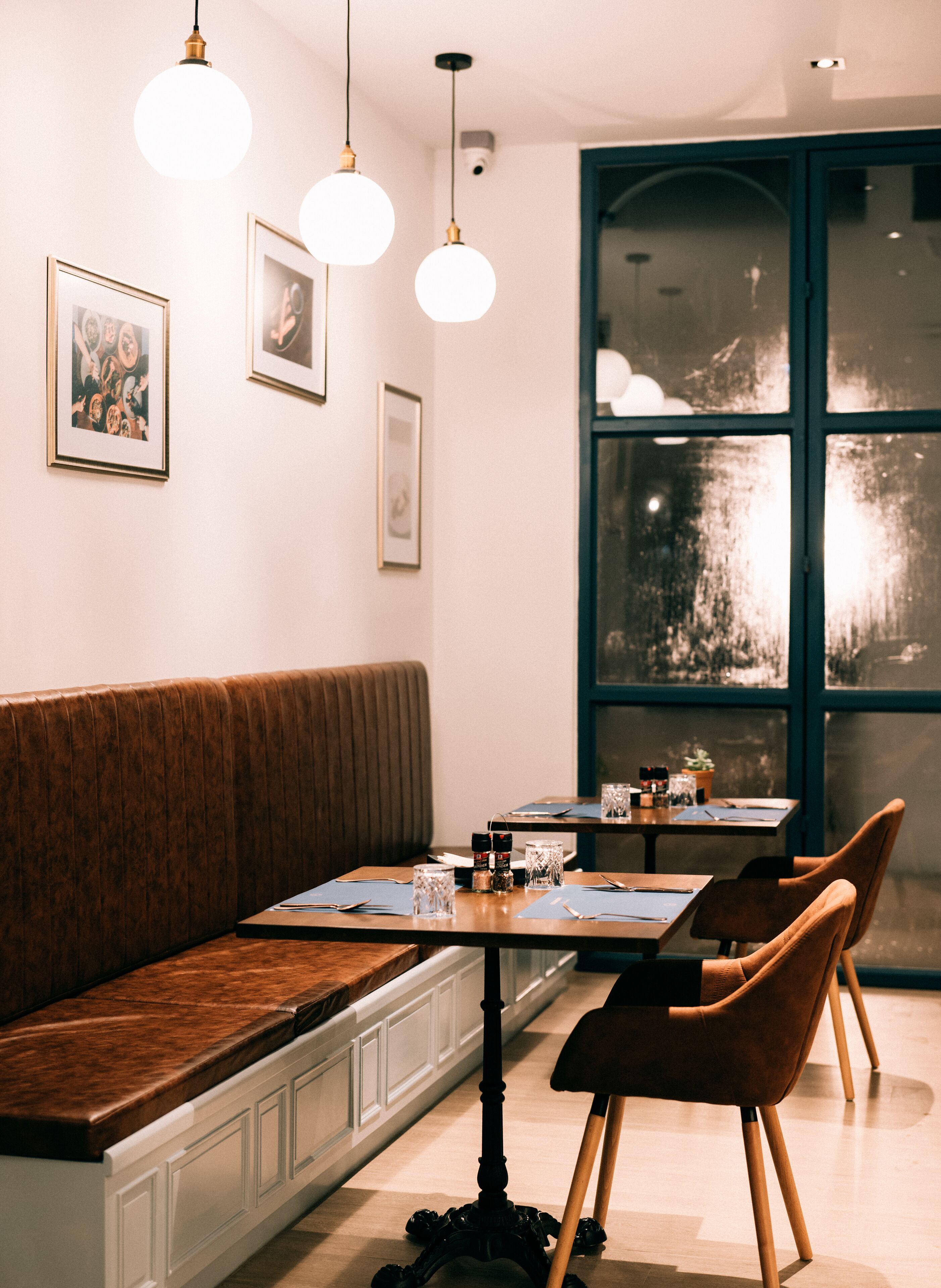 A restaurant with a wooden bench seating area, tables with chairs, pendant lighting, framed pictures on the wall, and a large window with a view.