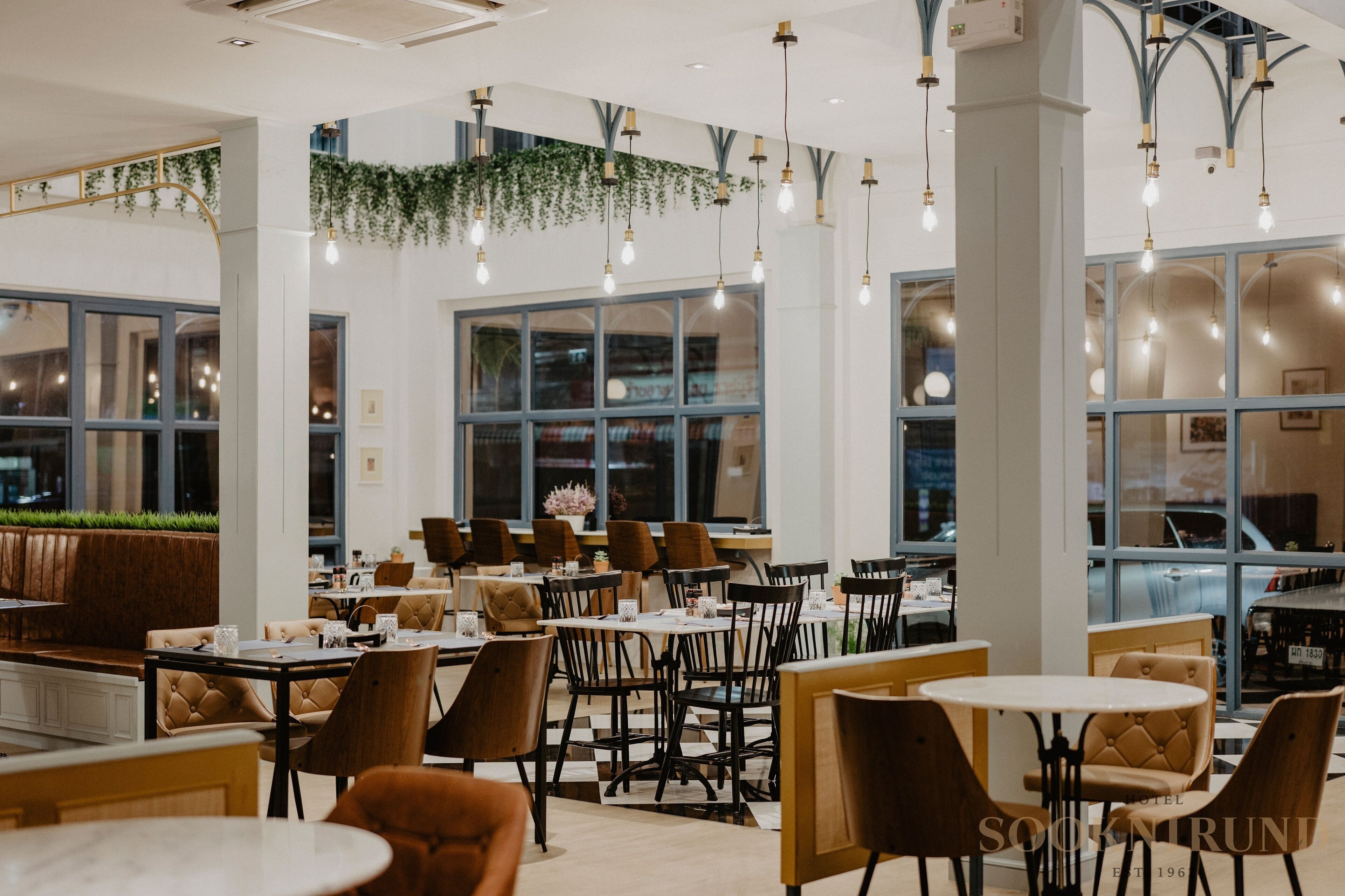 A modern restaurant with high ceilings, hanging light fixtures, and large windows. Tables and chairs are arranged for dining.