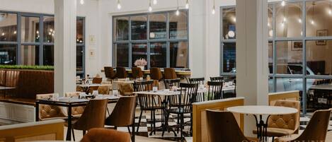 A modern restaurant with high ceilings, hanging light fixtures, and large windows. Tables and chairs are arranged for dining.