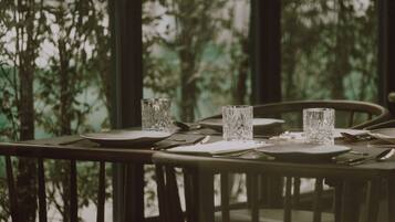 A dining table set with plates, cutlery, and glasses, with a view of trees through large windows.