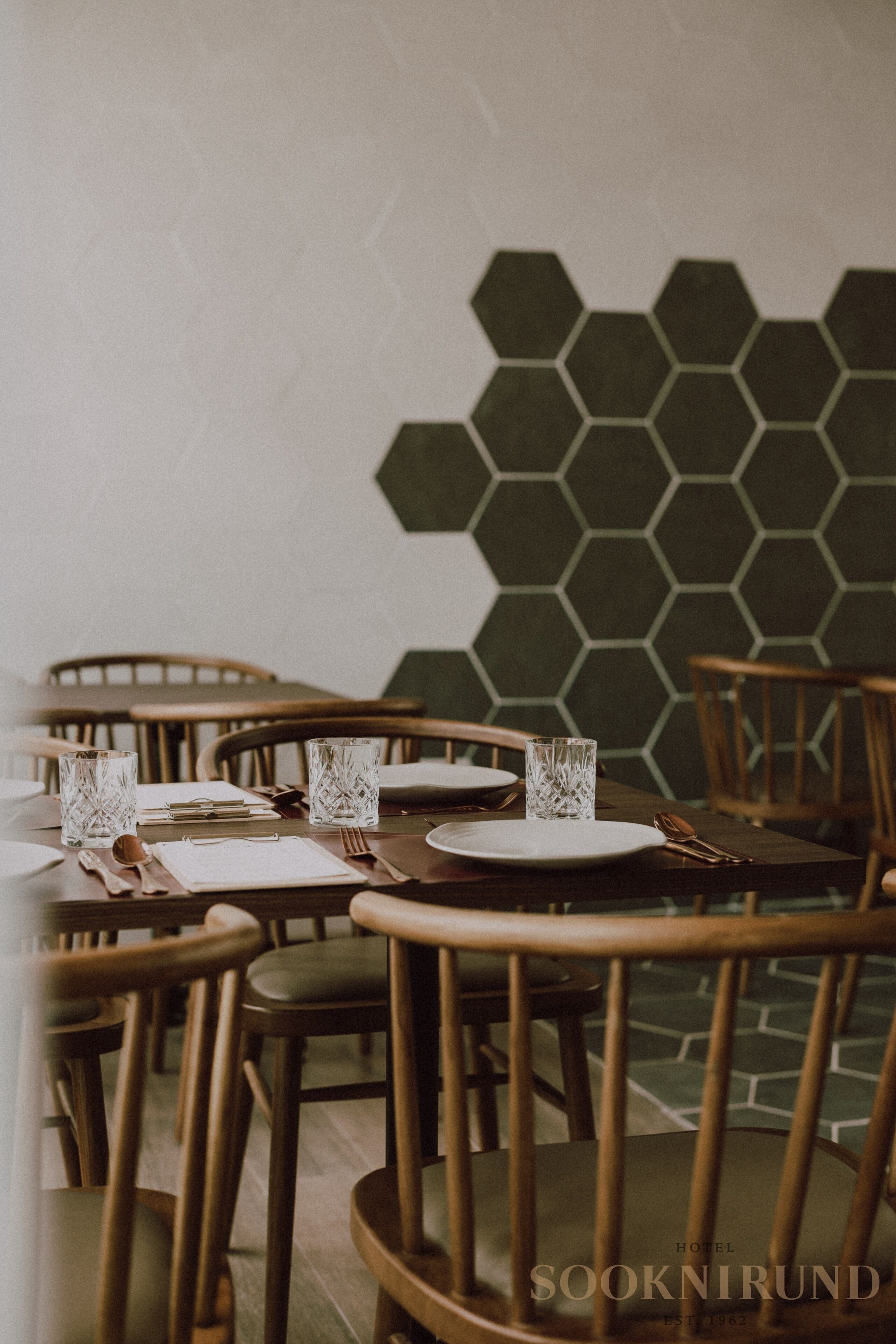 Comedor con pared de diseño hexagonal, sillas de madera y mesas dispuestas con cubiertos y vasos.