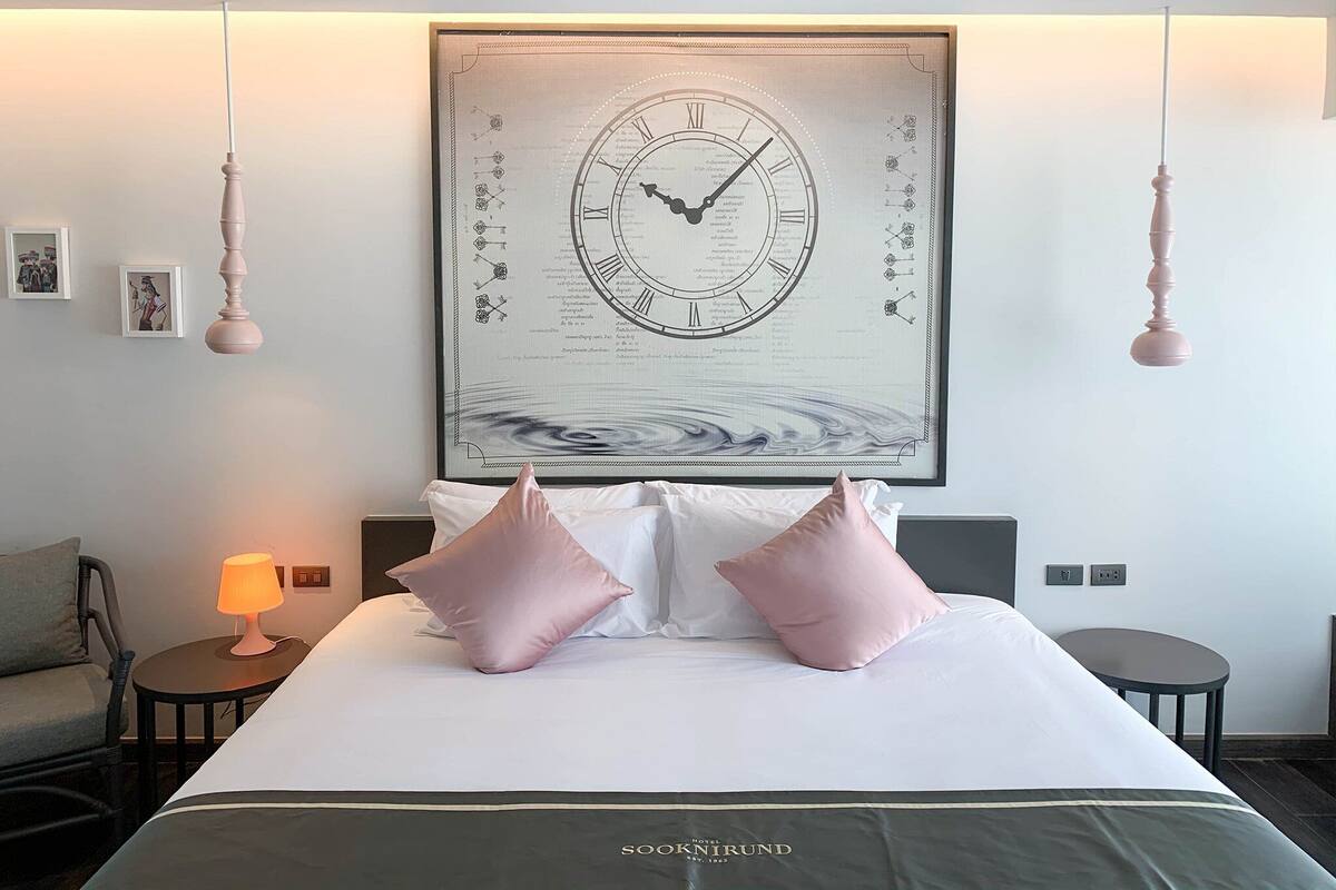 A hotel room with a large wall clock, a bed with white and pink pillows, a bedside table with a lamp, and a small round table.