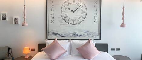 A hotel room with a large wall clock, a bed with white and pink pillows, a bedside table with a lamp, and a small round table.