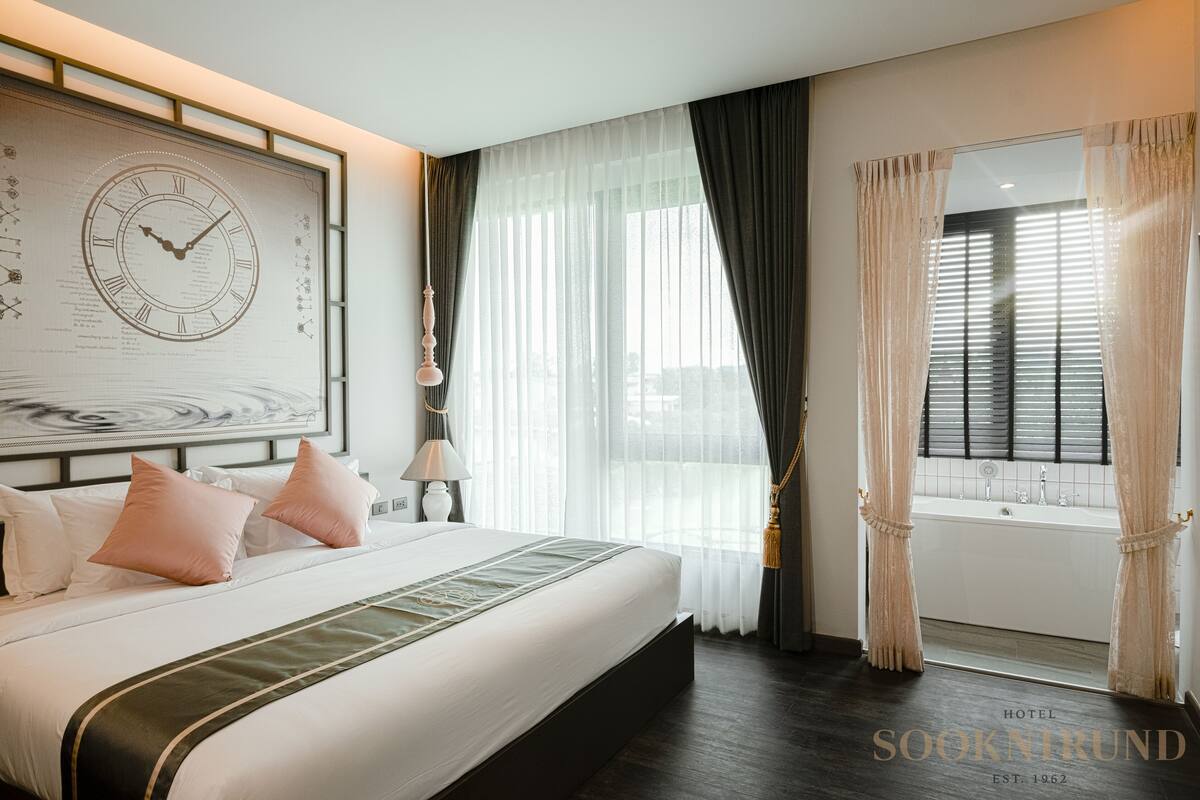 A hotel room with a large bed, a wall clock, a window with curtains, and a bathtub.