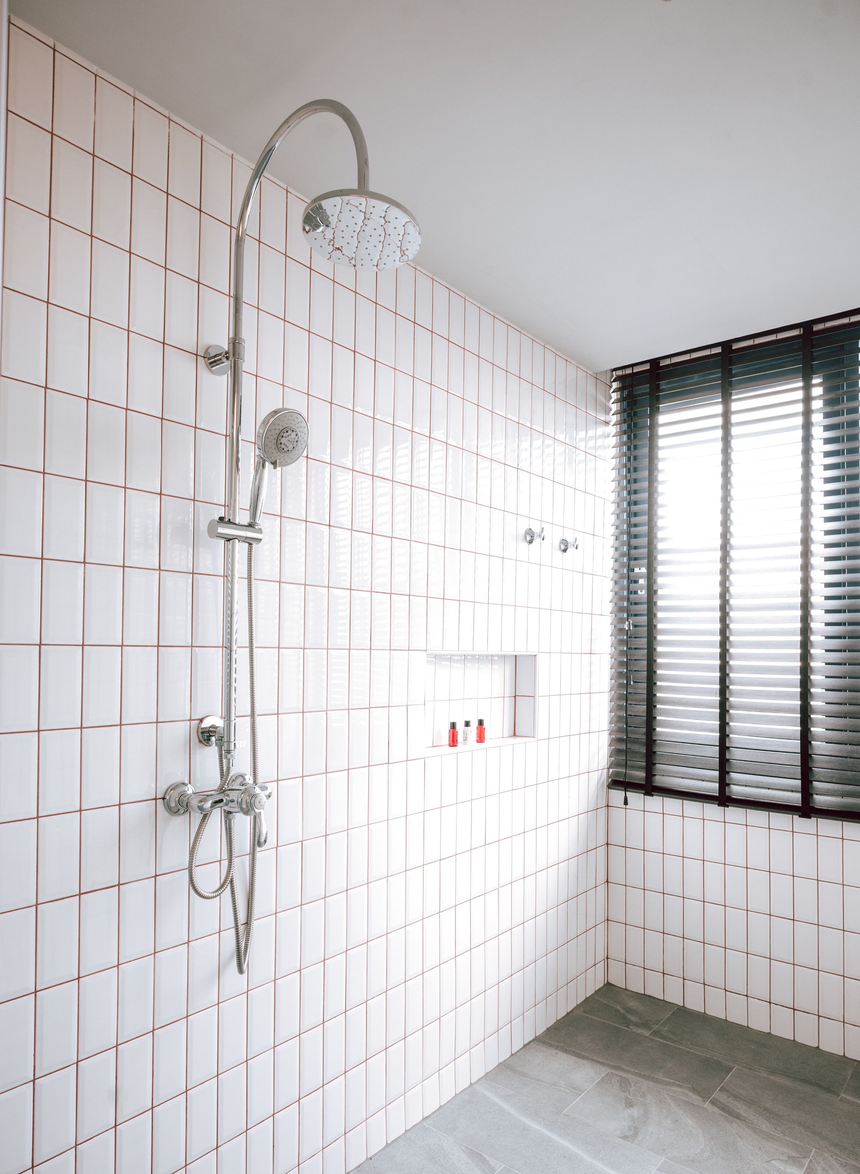 A bathroom with white tiled walls, a shower with a handheld showerhead, a glass-enclosed medicine cabinet, and a window with blinds.
