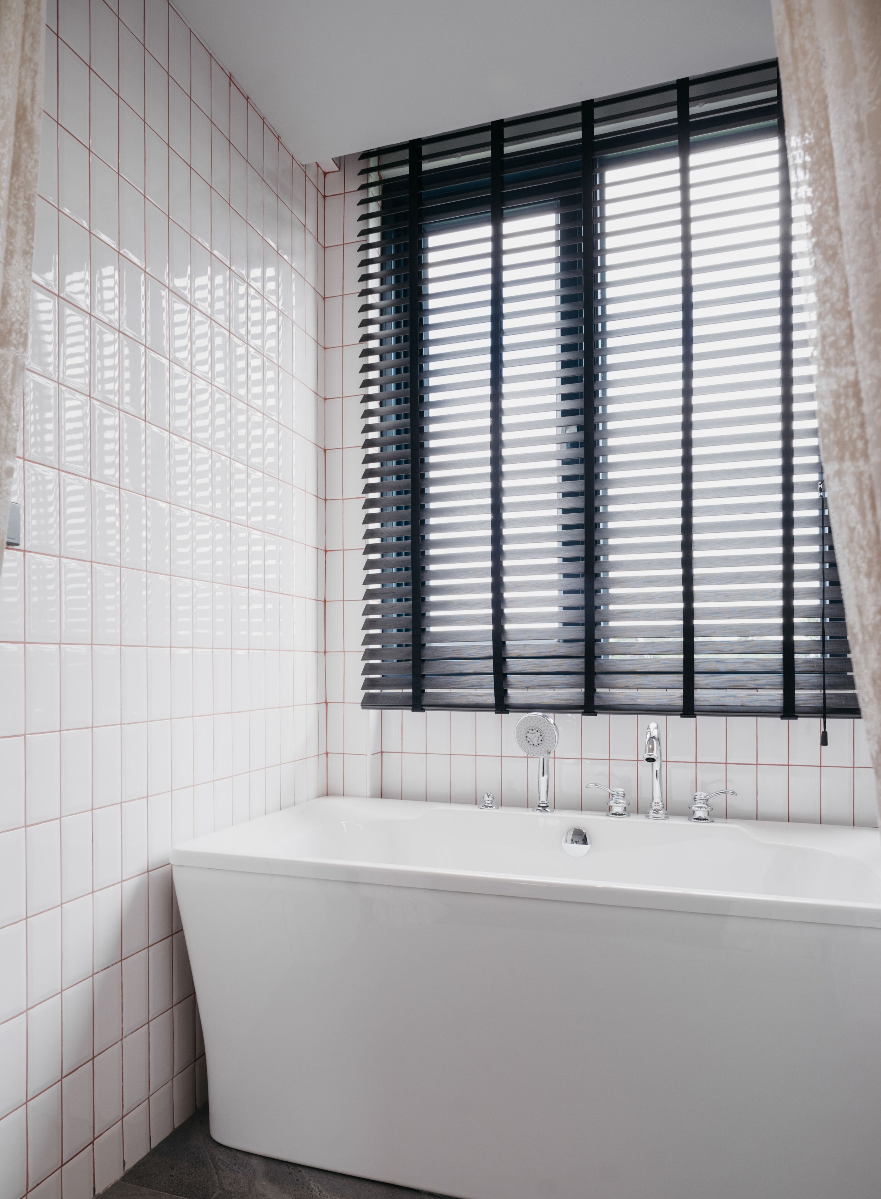 A bathroom with a white bathtub, a window with black blinds, and a tiled wall.
