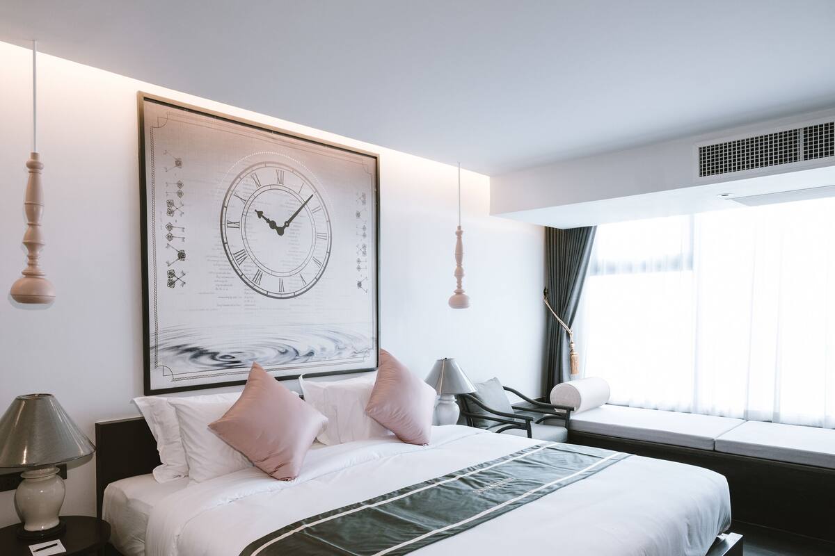 A modern hotel room with a large wall clock, a bed with white and pink bedding, a side table with a lamp, and a window with curtains.