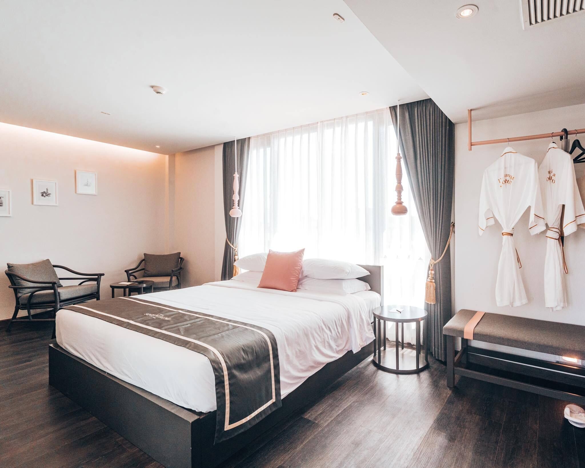 A modern hotel room with a large bed, bedside tables, a seating area with chairs, and a wardrobe with robes.