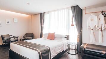 A modern hotel room with a large bed, bedside tables, a seating area with chairs, and a wardrobe with robes.