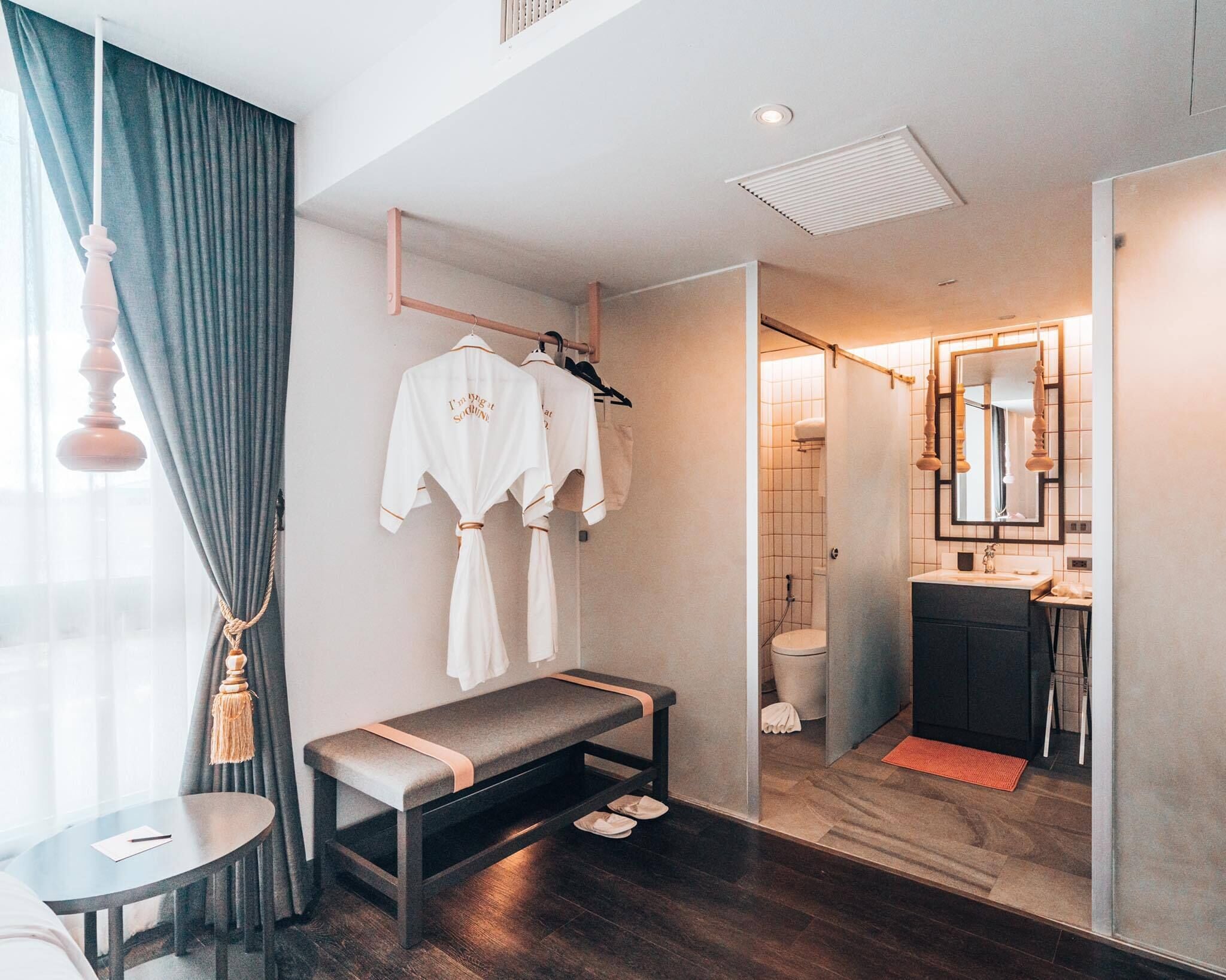 A modern hotel room with a bench, a table, a bench with shoes, a shower area, and a bathroom with a mirror and sink.