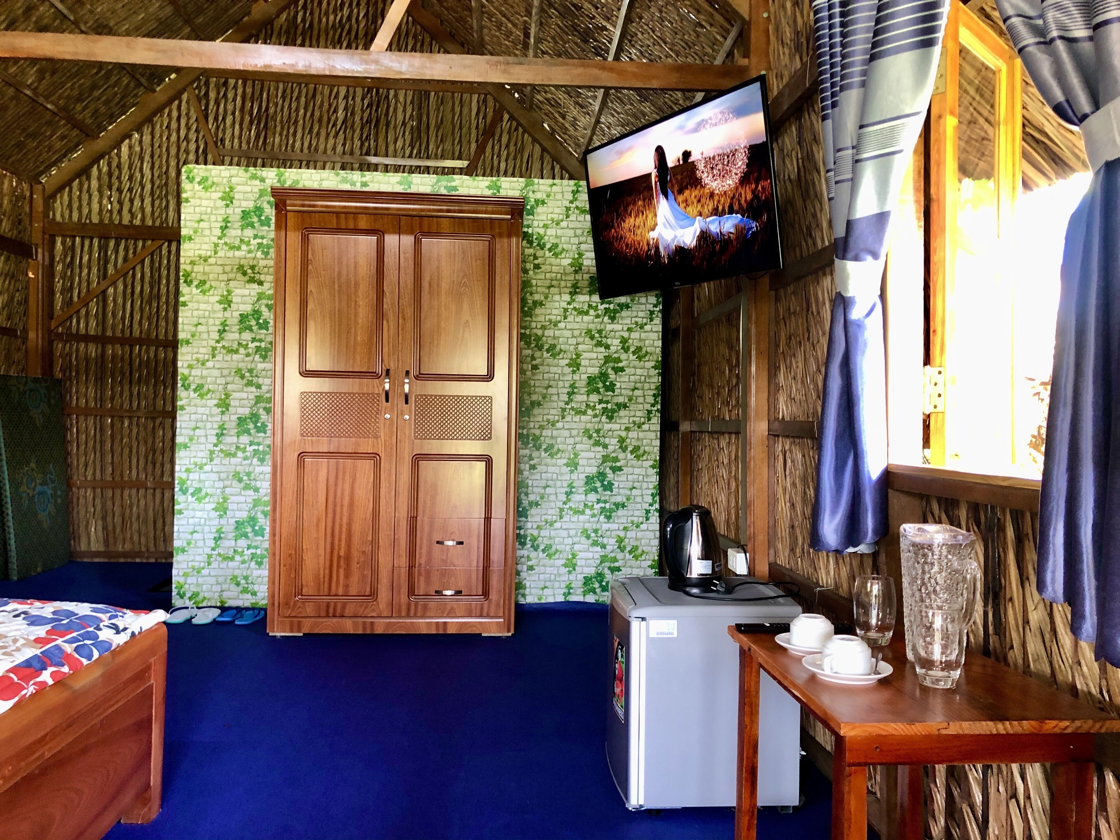 A room with a wooden wardrobe, a television mounted on the wall, a small refrigerator, and a table with cups and a glass.