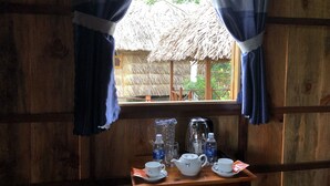 Una habitación de madera con ventana que da a una edificación con techo de paja. En la habitación, una mesa tiene un hervidor, tazas y agua embotellada.